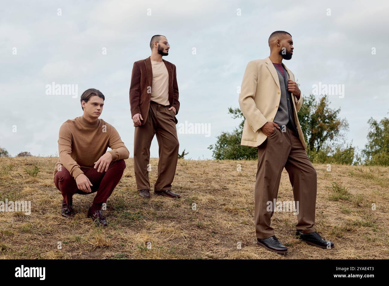 Drei gutaussehende Männer in trendiger Herbstkleidung posieren selbstbewusst auf einem ruhigen Feld unter bewölktem Himmel. Stockfoto