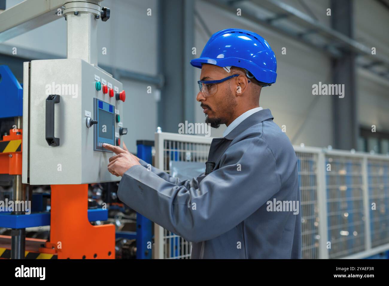 Afroamerikanischer Ingenieur, der einen grauen Arbeitsmantel und einen blauen Helm trägt und die Produktion und Programmierung einer Maschine in einer Fabrik steuert. Stockfoto