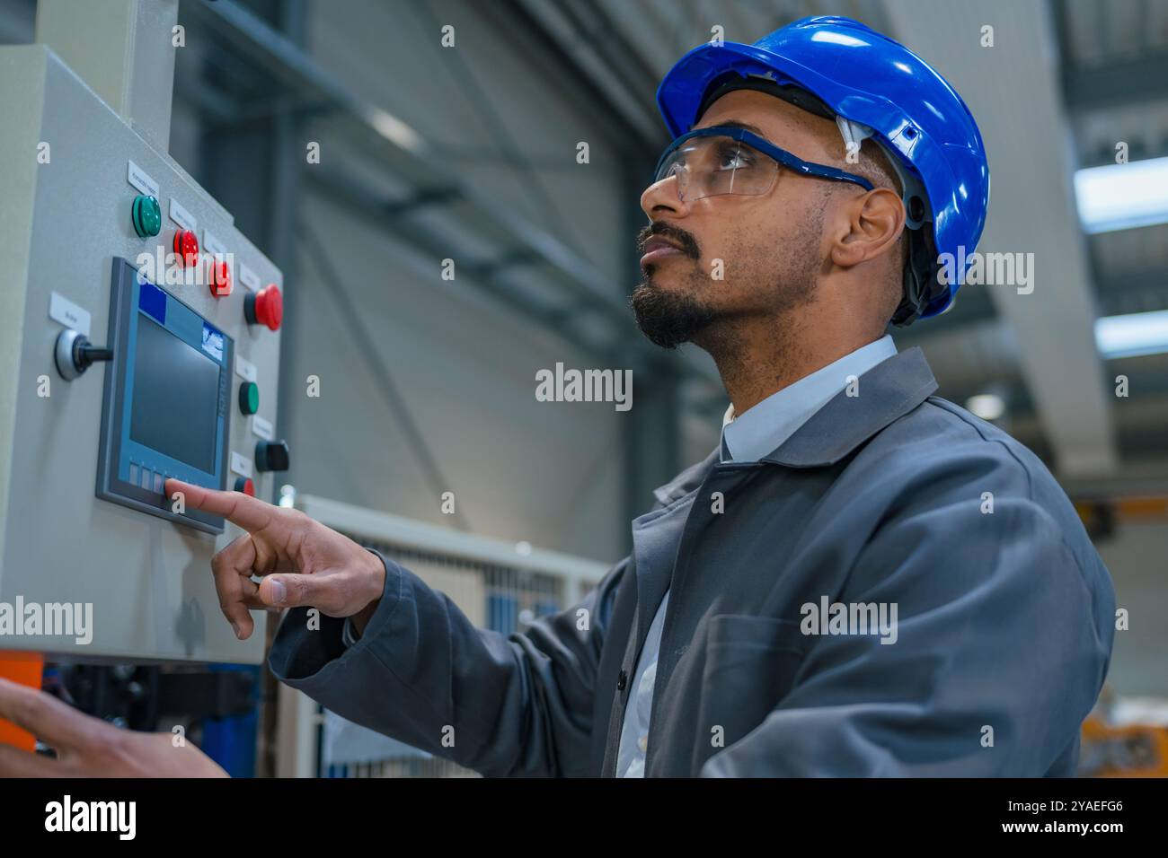 Afroamerikanischer Ingenieur, der einen grauen Arbeitsmantel und einen blauen Helm trägt und die Produktion und Programmierung einer Maschine in einer Fabrik steuert. Stockfoto