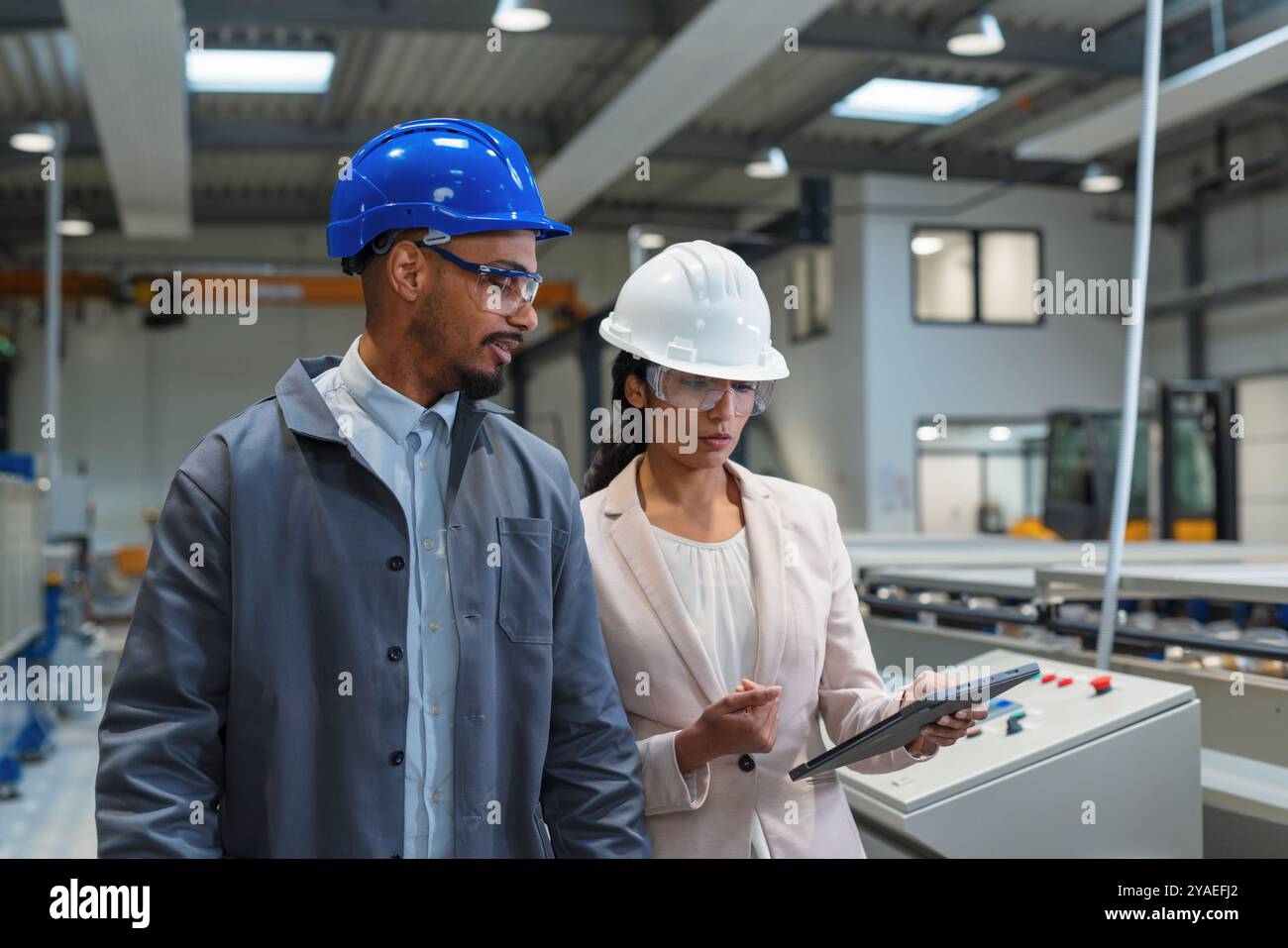 Afroamerikaner Ingenieur und eine Inderin, eine Fabrikleiterin, die ein Tablet hält, durch die Produktionslinie läuft, Inspektionen durchführt, Vorderansicht. Stockfoto