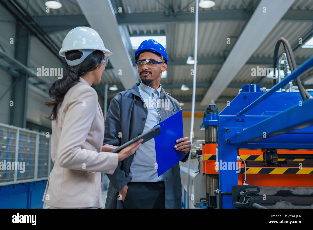 Afroamerikaner Ingenieur und eine Inderin, eine Fabrikleiterin, die ein Tablet hält, durch die Produktionslinie läuft, Inspektionen durchführt, Vorderansicht. Stockfoto