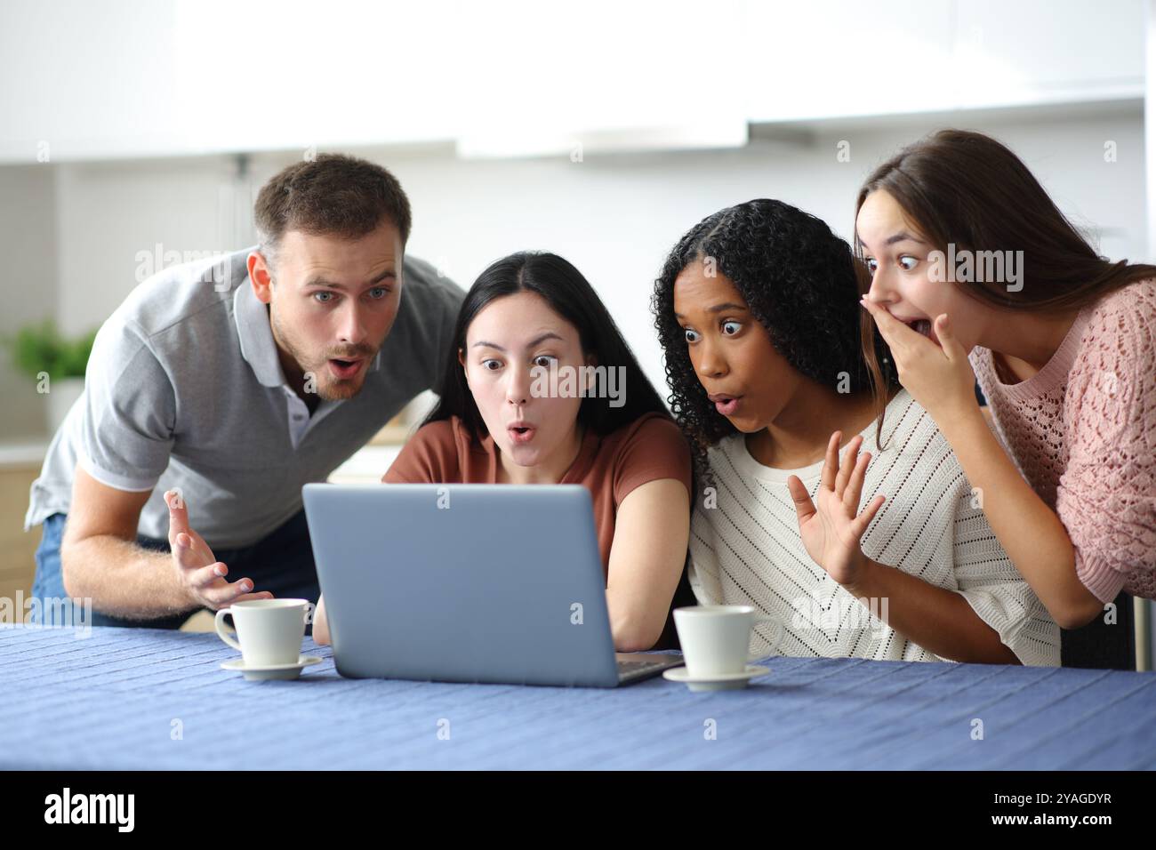 Vier verblüffte Mitbewohner, die in der Küche zu Hause überraschende Nachrichten auf dem Laptop lesen Stockfoto