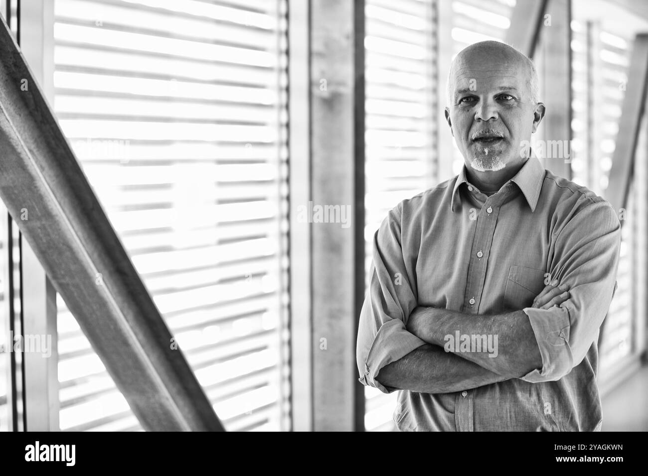 Schwarz-weiß-Porträt eines selbstbewussten IT-Managers, der mit gekreuzten Armen in einem modernen Bürogebäude steht und Erfahrung und Führungskräfte vermittelt Stockfoto