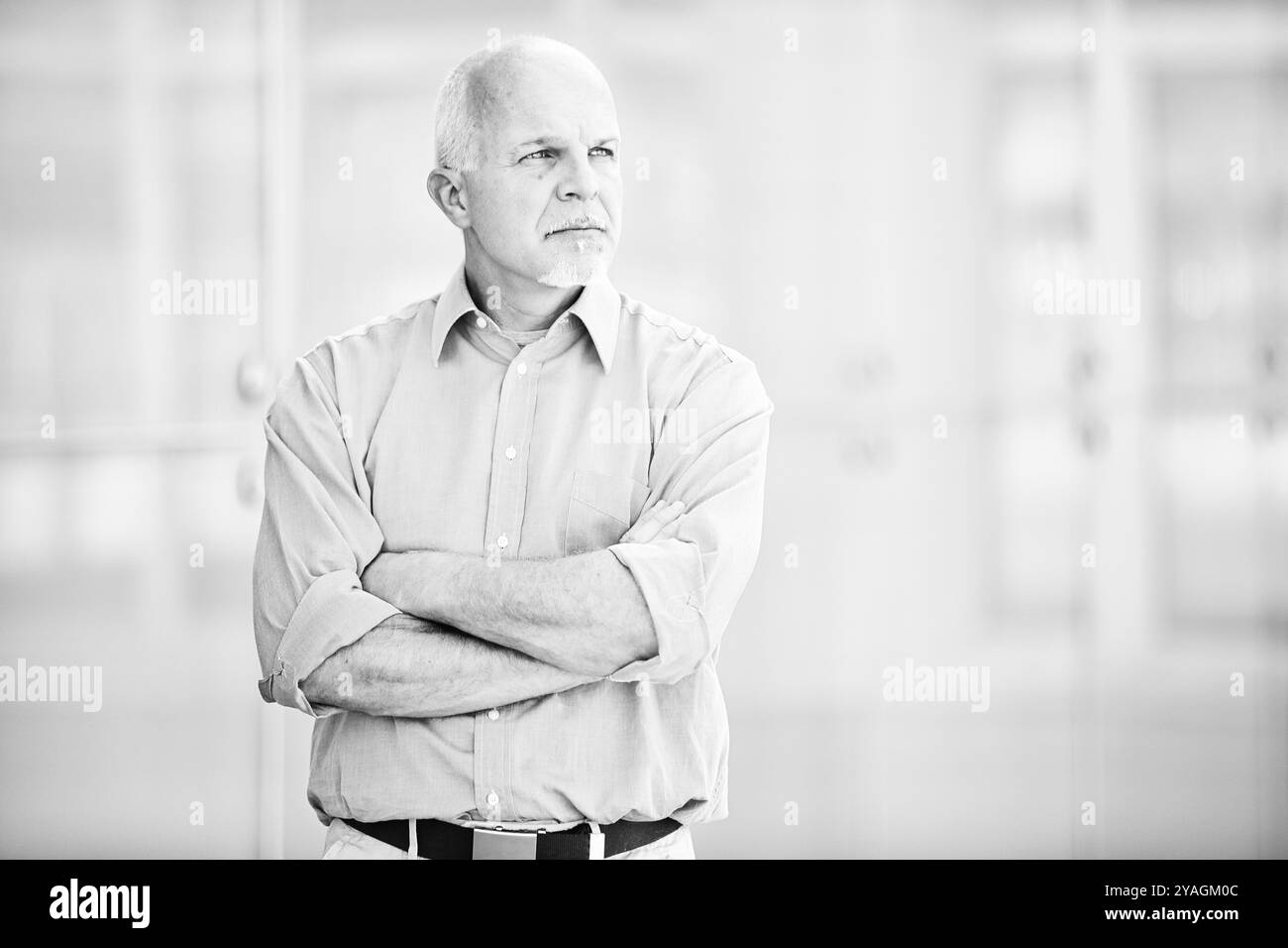 Schwarz-weiß-Porträt eines leitenden Managers, der mit überkreuzten Armen in einem modernen Bürogebäude steht und besinnlich von der Kamera wegblickt Stockfoto