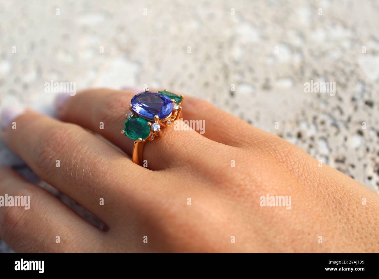 Hand trägt einen goldenen Ring mit einem großen blauen Edelstein in der Mitte, flankiert von zwei kleineren grünen Edelsteinen und mit kleinen Diamanten, besetzt mit Agave Stockfoto