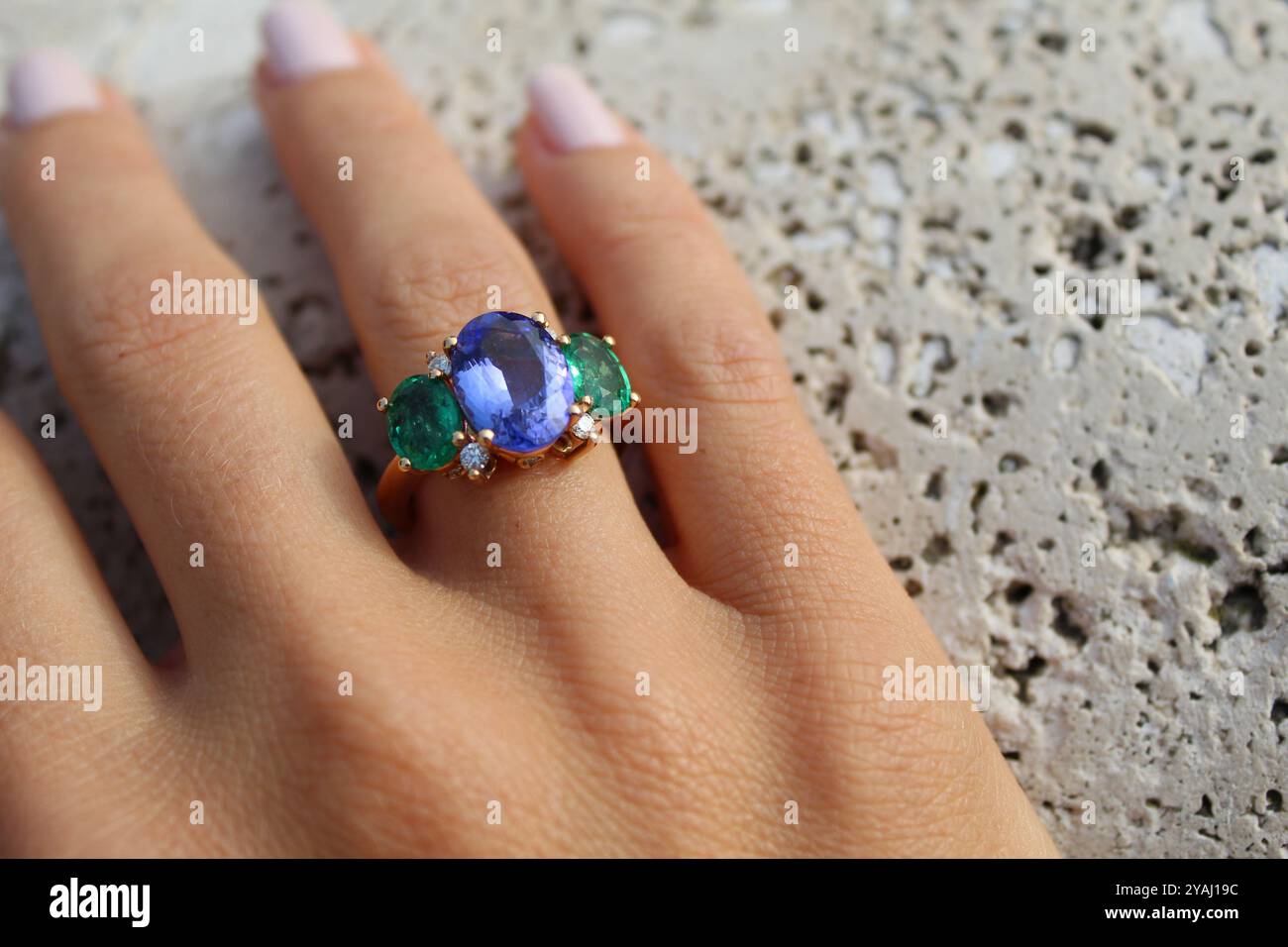 Hand trägt einen goldenen Ring mit einem großen blauen Edelstein in der Mitte, flankiert von zwei kleineren grünen Edelsteinen und mit kleinen Diamanten, besetzt mit Agave Stockfoto