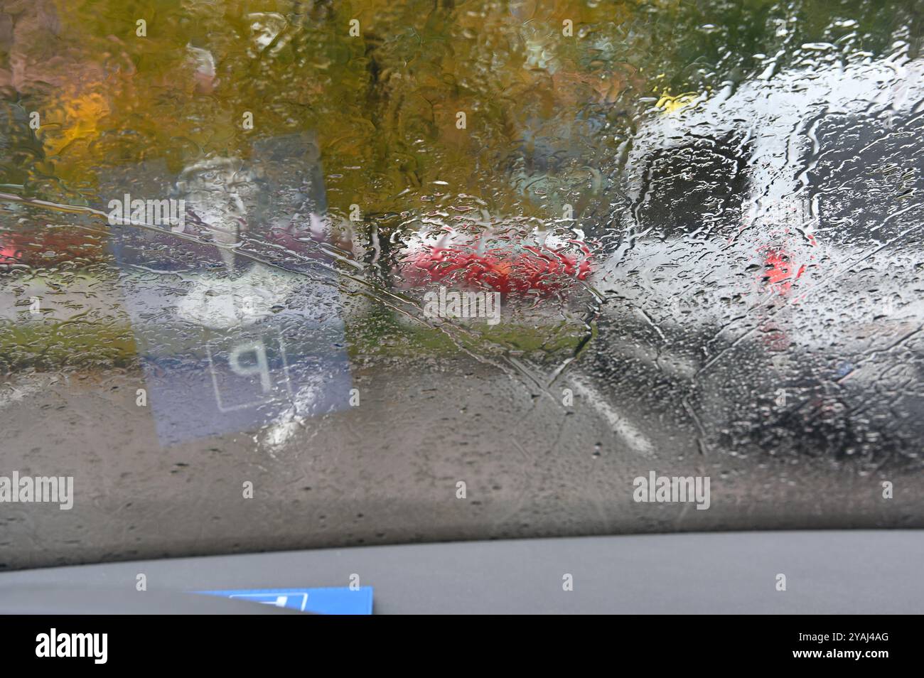 Das Foto zeigt eine regnerische Tagesszene, die durch eine Windschutzscheibe des Autos gesehen wird. Die Wassertropfen und -Streifen des Regens verzerren die Sicht nach draußen und machen es so Stockfoto