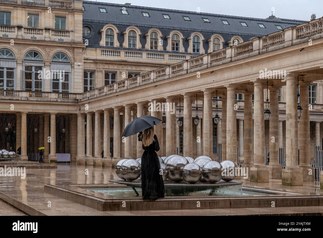 Eine blonde Frau in schwarz gekleidet mit einem schwarzen Schirm vor dem alten Palais-Royal in Paris, Frankreich Stockfoto