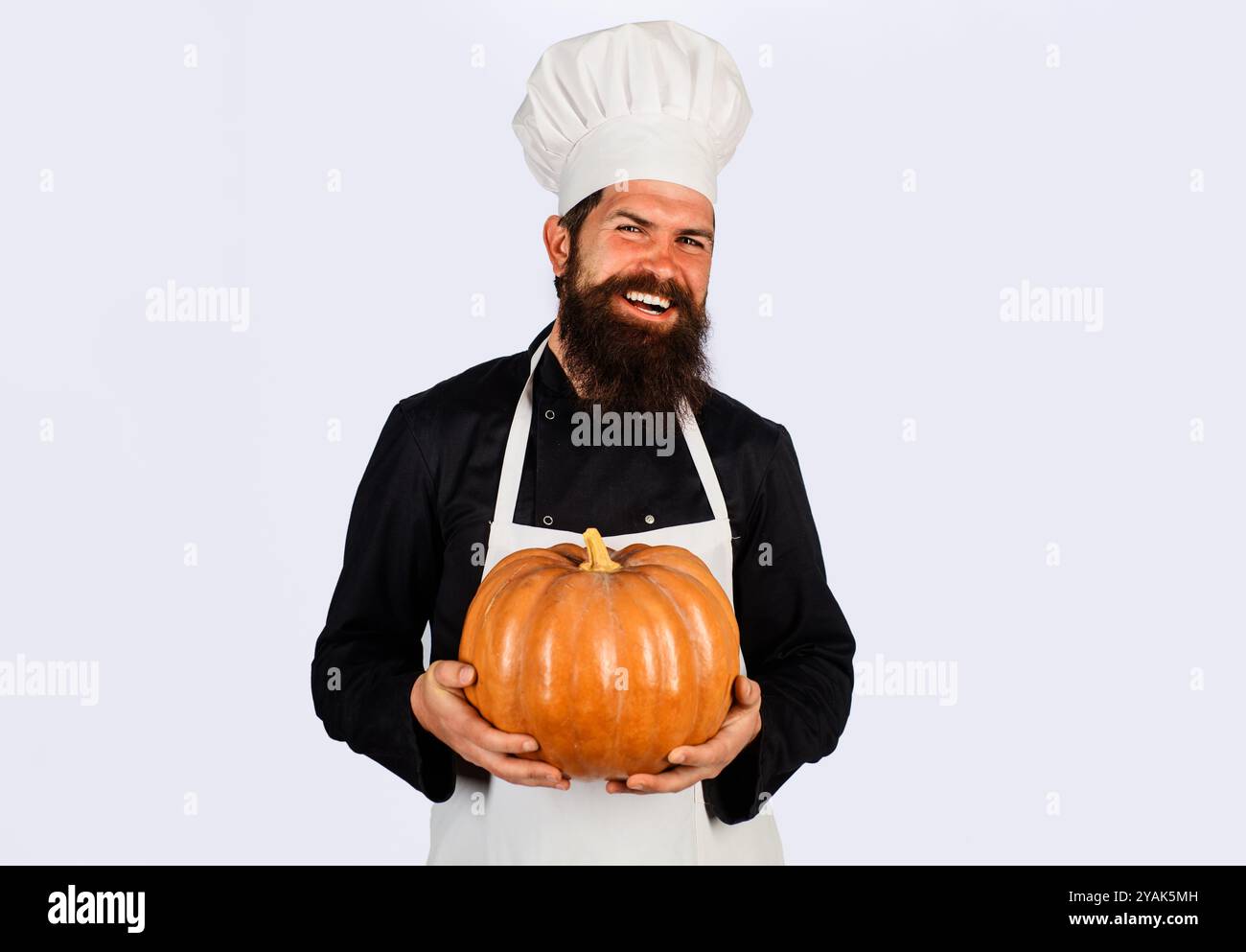 Lächelnder männlicher Koch in Kochmütze mit Kürbis. Gesunde vegetarische Ernährung. Bauernmarkt. Herbstrezepte. Bärtiger Mann in Küchenuniform mit Kürbis für Hallo Stockfoto