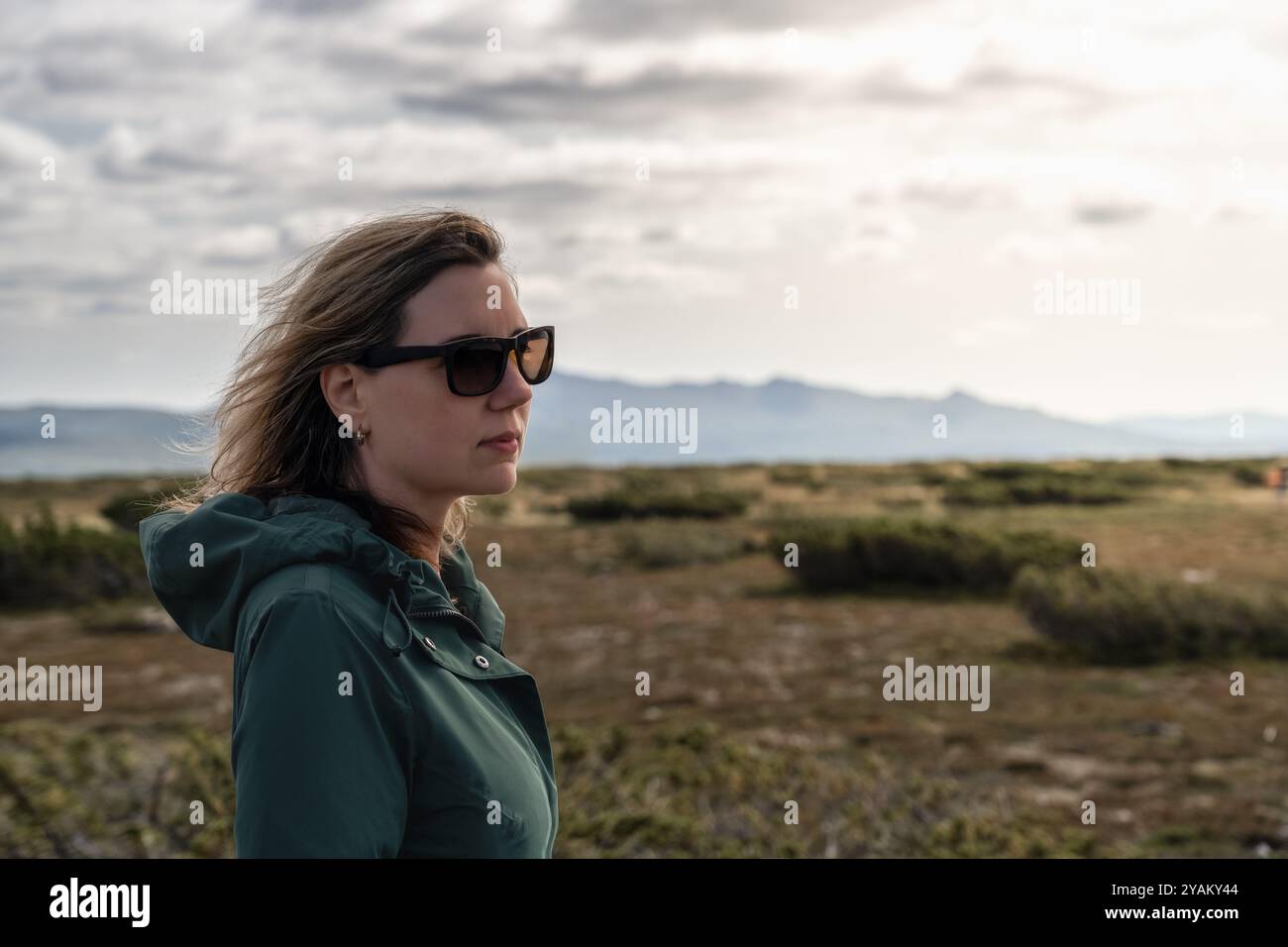 Frau in Sonnenbrille steht in der weitläufigen Landschaft, mit Blick auf majestätische Berge, sanft im Wind wehenden Haaren, hell glühendes Sonnenlicht auf ihrem Gesicht Stockfoto