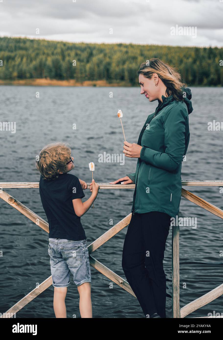 Mutter und Sohn sehen sich fröhlich an, während sie geröstete Marshmallows draußen an einem ruhigen See essen, was einen herzerwärmenden Moment in der Natur schafft Stockfoto