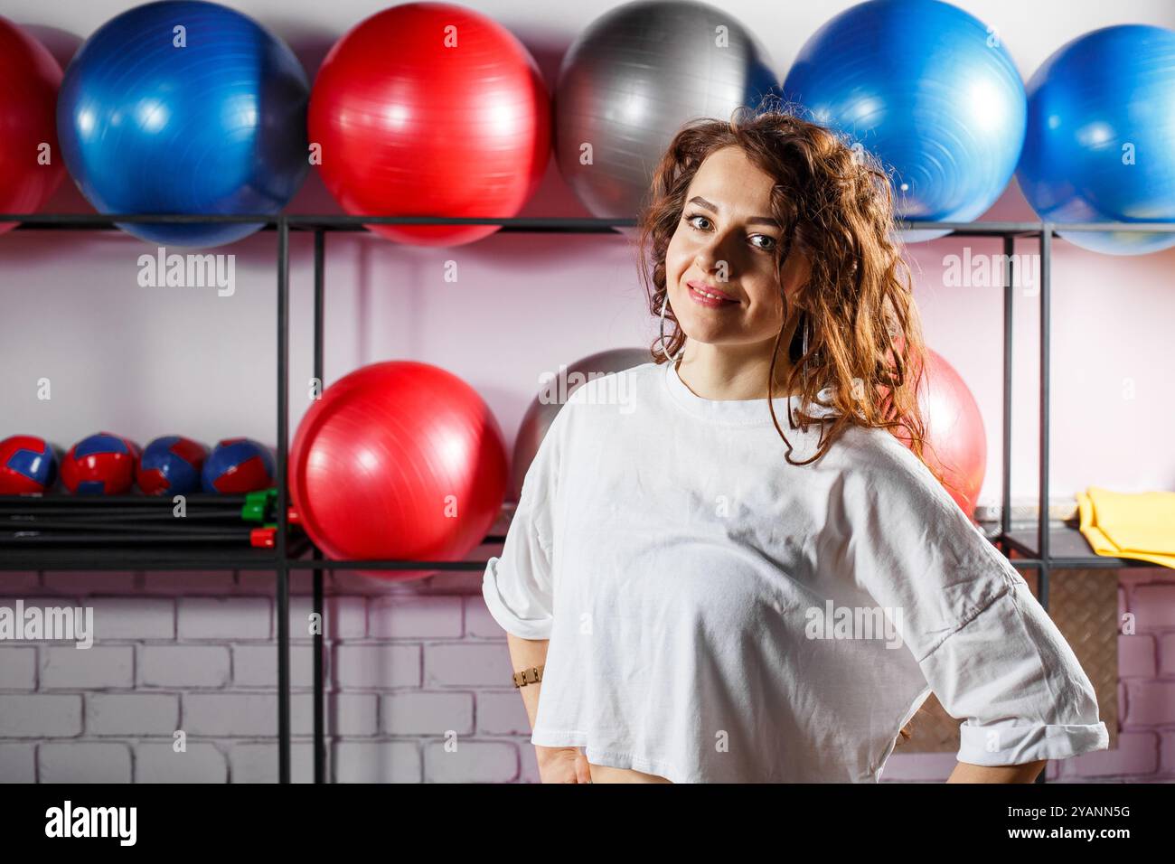 Eine junge Frau mit lockigen Haaren steht lächelnd in einem Fitnessstudio. Hinter ihr befinden sich lebendige Übungsbälle und Gewichte in den Regalen. Stockfoto