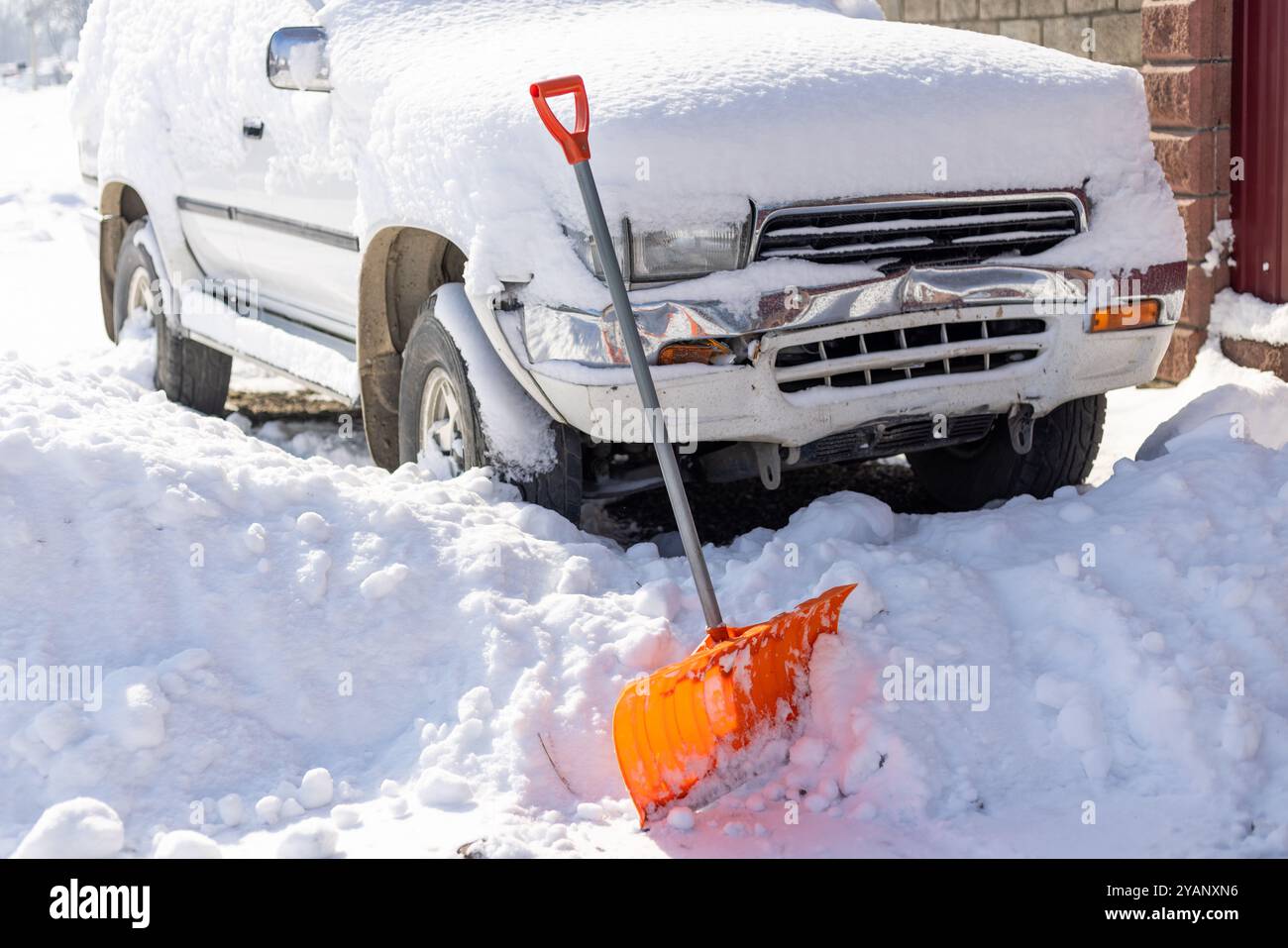 Die orangefarbene Schneeschaufel ragt aus der Schneewolke vor dem schneebedeckten weißen SUV-Auto hervor Stockfoto