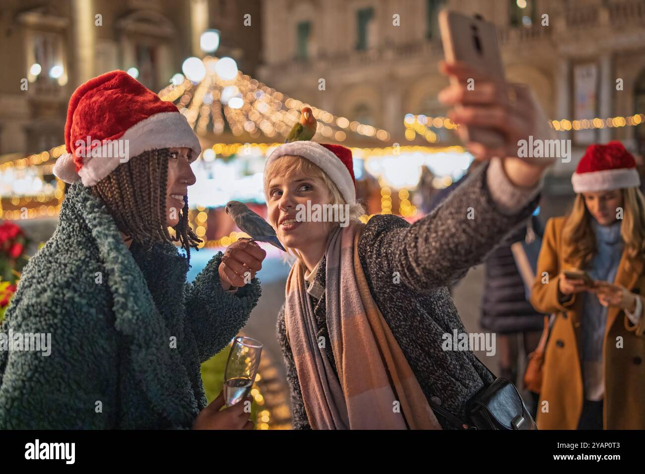 Verschiedene Freunde machen Selfie auf dem Weihnachtsmarkt. Frauen mit Weihnachtsmützen lächeln und posieren mit Sektgläsern, umgeben von festlichen Lichtern Stockfoto