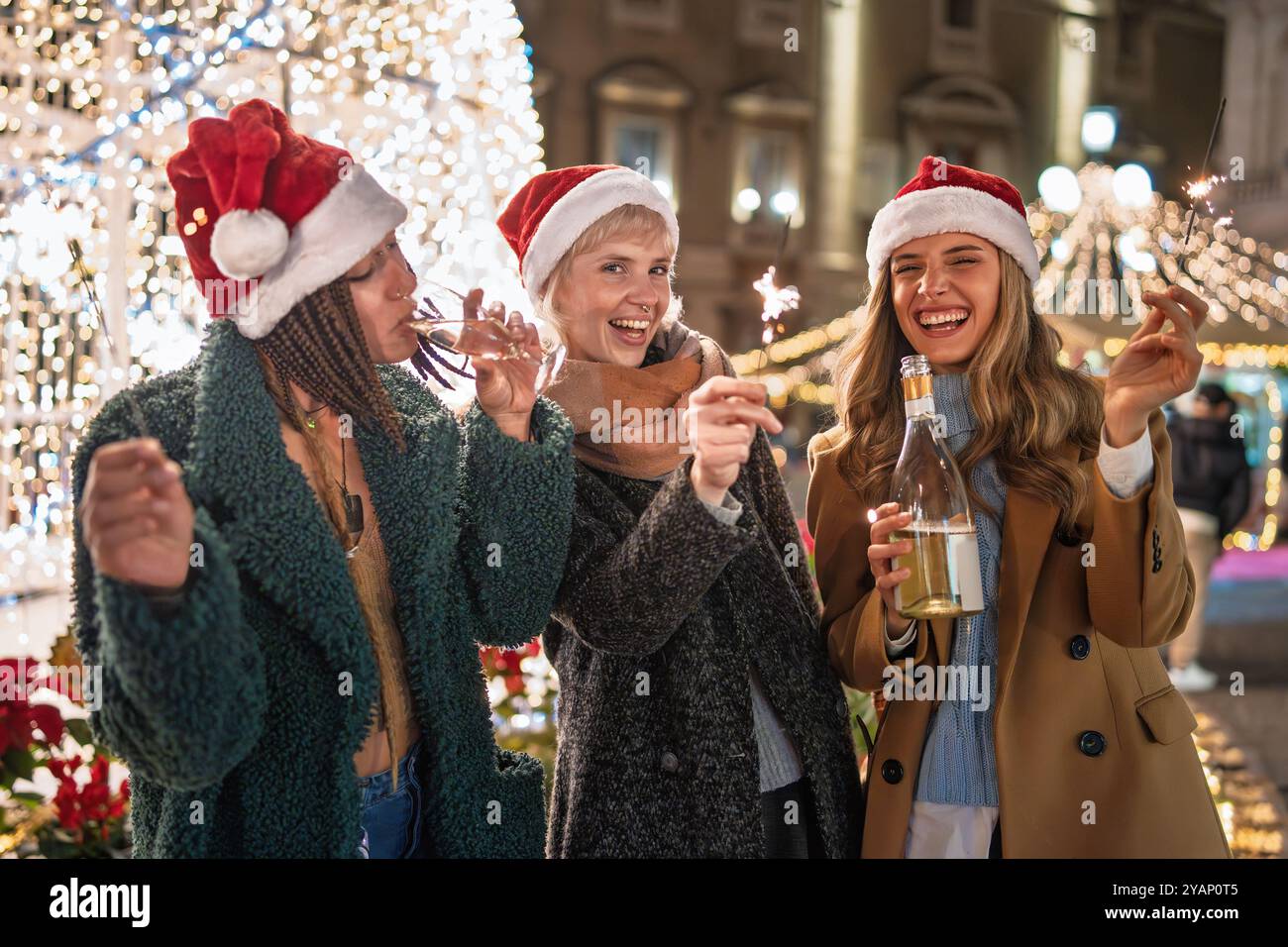 Verschiedene Gruppen junger Frauen feiern Weihnachten auf einem Festmarkt. Freunde in Santa Hüte lachen und genießen Sparkler und Getränke umgeben von h Stockfoto