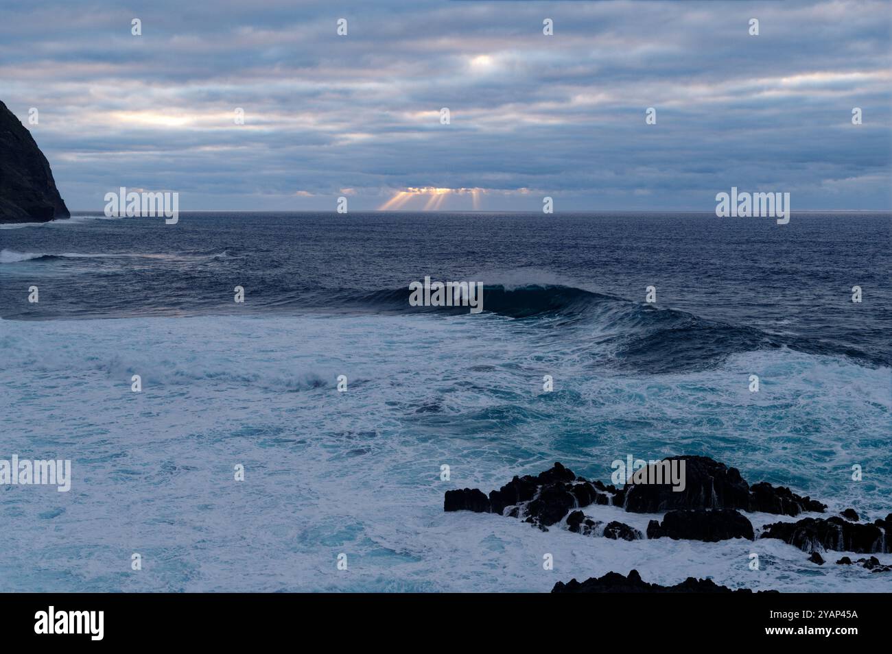 Sonnenstrahlen brechen durch die Wolken über dem atlantik und werfen ein ruhiges Licht auf die Wellen vor Madeiras dramatischer Küste Stockfoto