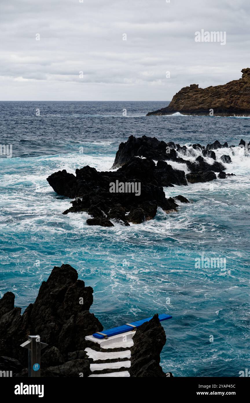 Wellen schlagen gegen die vulkanischen Felsen und ein blaues Tauchbrett an den natürlichen Pools von Porto Moniz Stockfoto