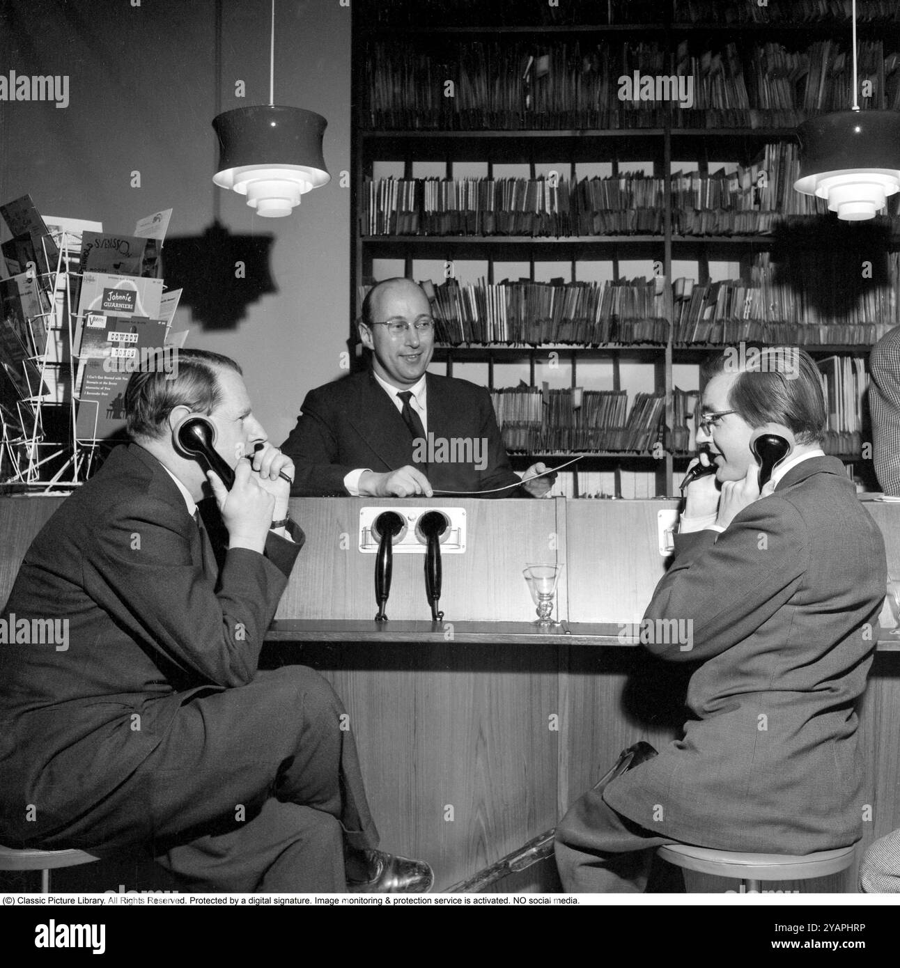 Wie Sie Platten in den 1950ern gekauft haben Die Männer, die ein Telefon halten, hören sich Aufzeichnungen an. Zu dieser Zeit war es üblich, dass die Kunden die Aufzeichnungen im Store anhören konnten, bevor sie sich für den Kauf entschieden. Der Plattenladen verfügt über mehrere Abhörstationen und die Mitarbeiter hinter dem Schalter, die das System verwalten. Dieses Geschäft ist die Selanders Skivbar auf der Vasagatan in Stockholm. 1955. Stockfoto