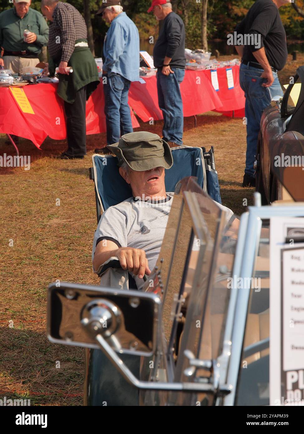 Ein älterer Mann, der auf einer Autoshow schläft, während er einen bequemen Hut trägt. Mann mit offenem Mund in einem Klappstuhl Stockfoto