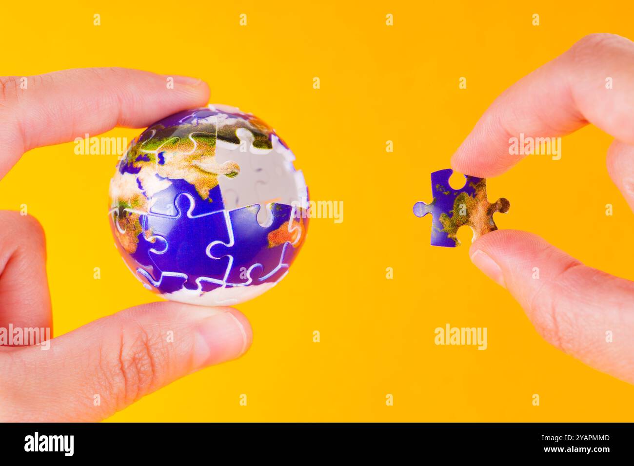Zwei Hände arbeiten mit einem Globus-Puzzleteil vor einem leuchtend gelben Hintergrund und heben Themen der globalen Verbindung und Kreativität hervor. Stockfoto