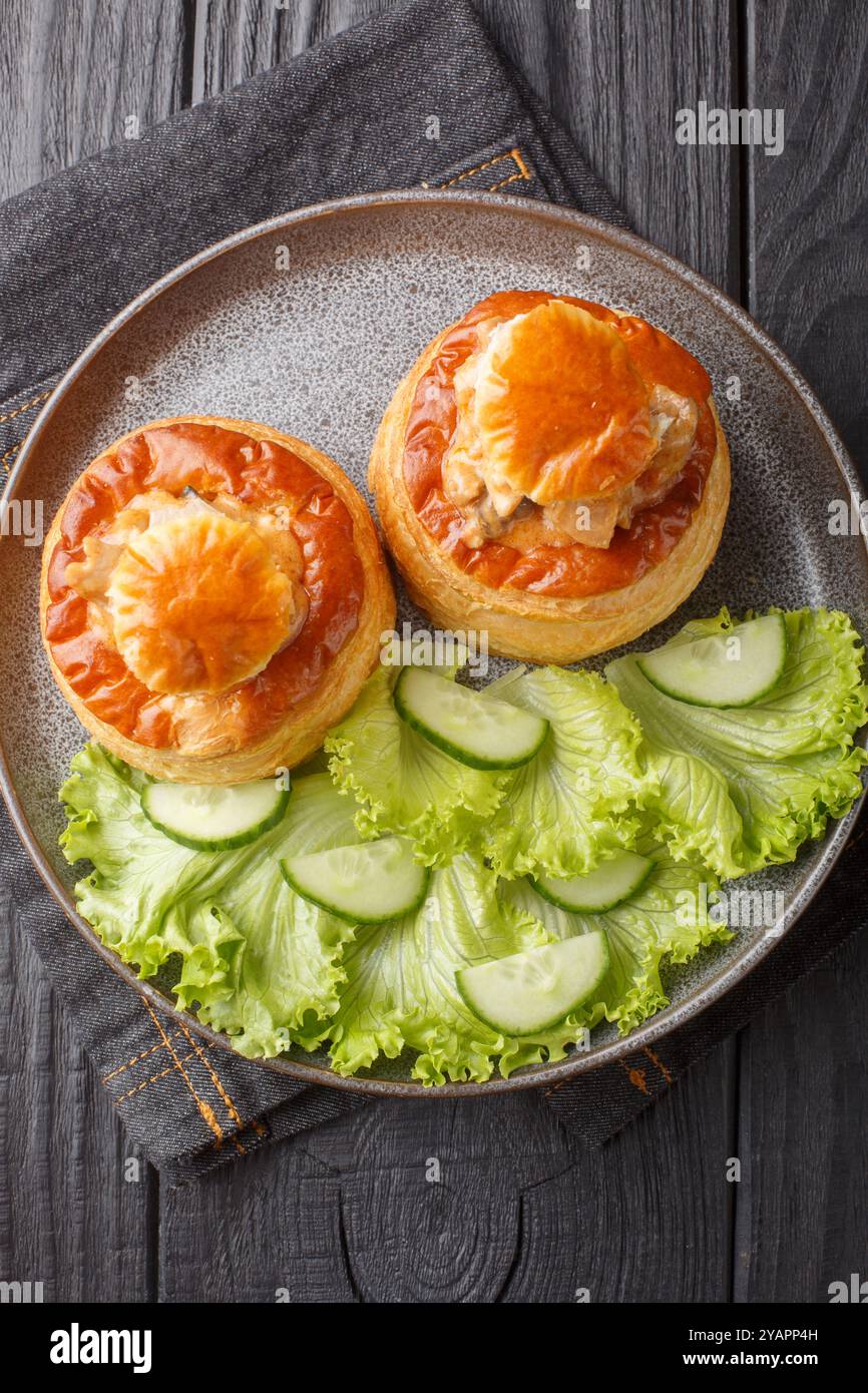 Bouchee a la reine oder vol-au-Vent mit Kalbfleisch in scharfer Sauce serviert mit Salat und Gurke in Nahaufnahme auf einem Teller auf einem Holztisch. Vertikale Draufsicht f Stockfoto