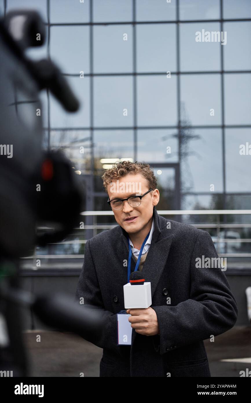 Ein Journalist mit Brille und Mantel führt ein Interview, während er ein Mikrofon vor einem Gebäude hält. Stockfoto