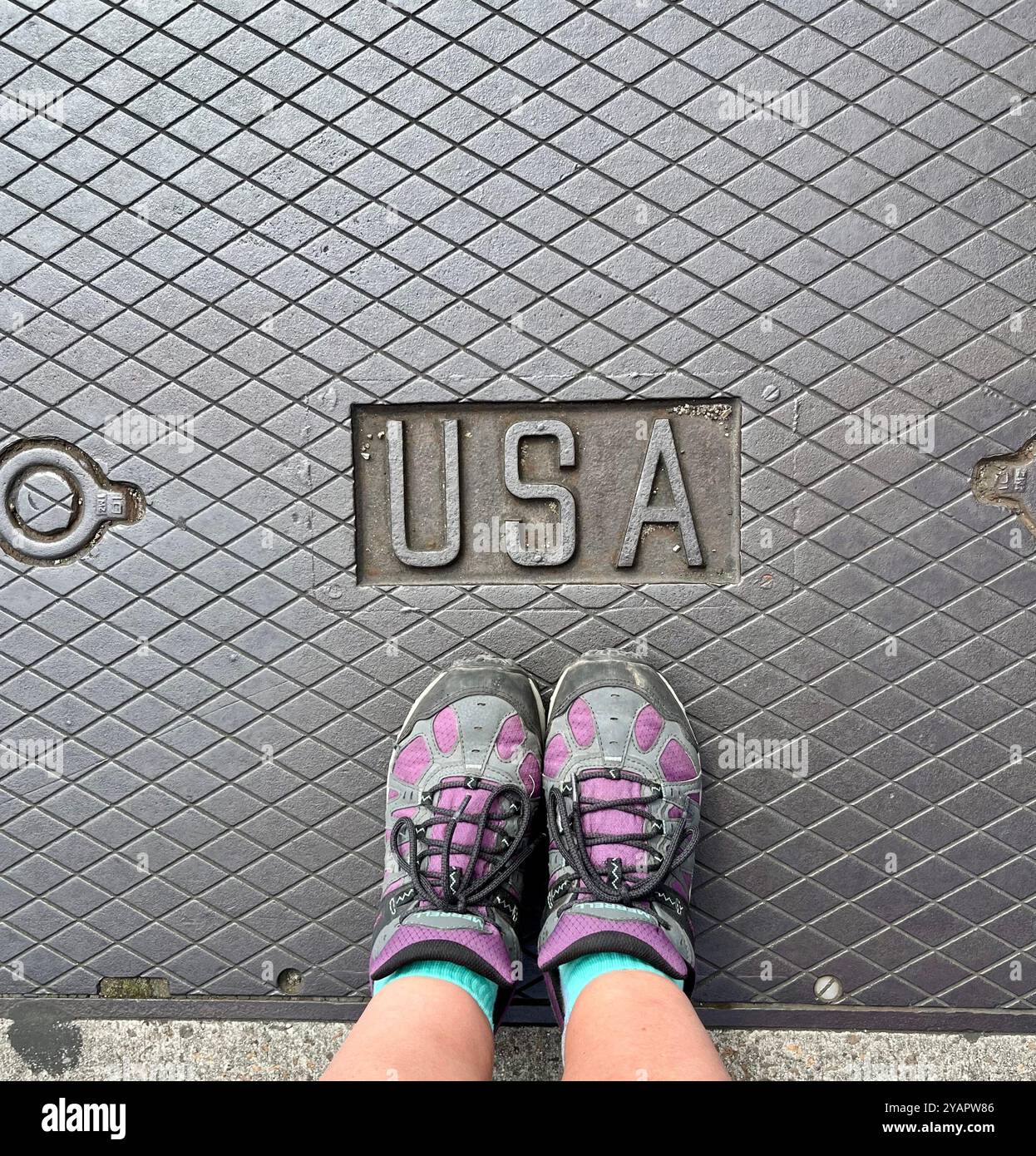 Füße in Merrell-Schuhen auf einem Eisenmann-Lochdeckel, auf dem die USA eingeprägt sind. Stockfoto