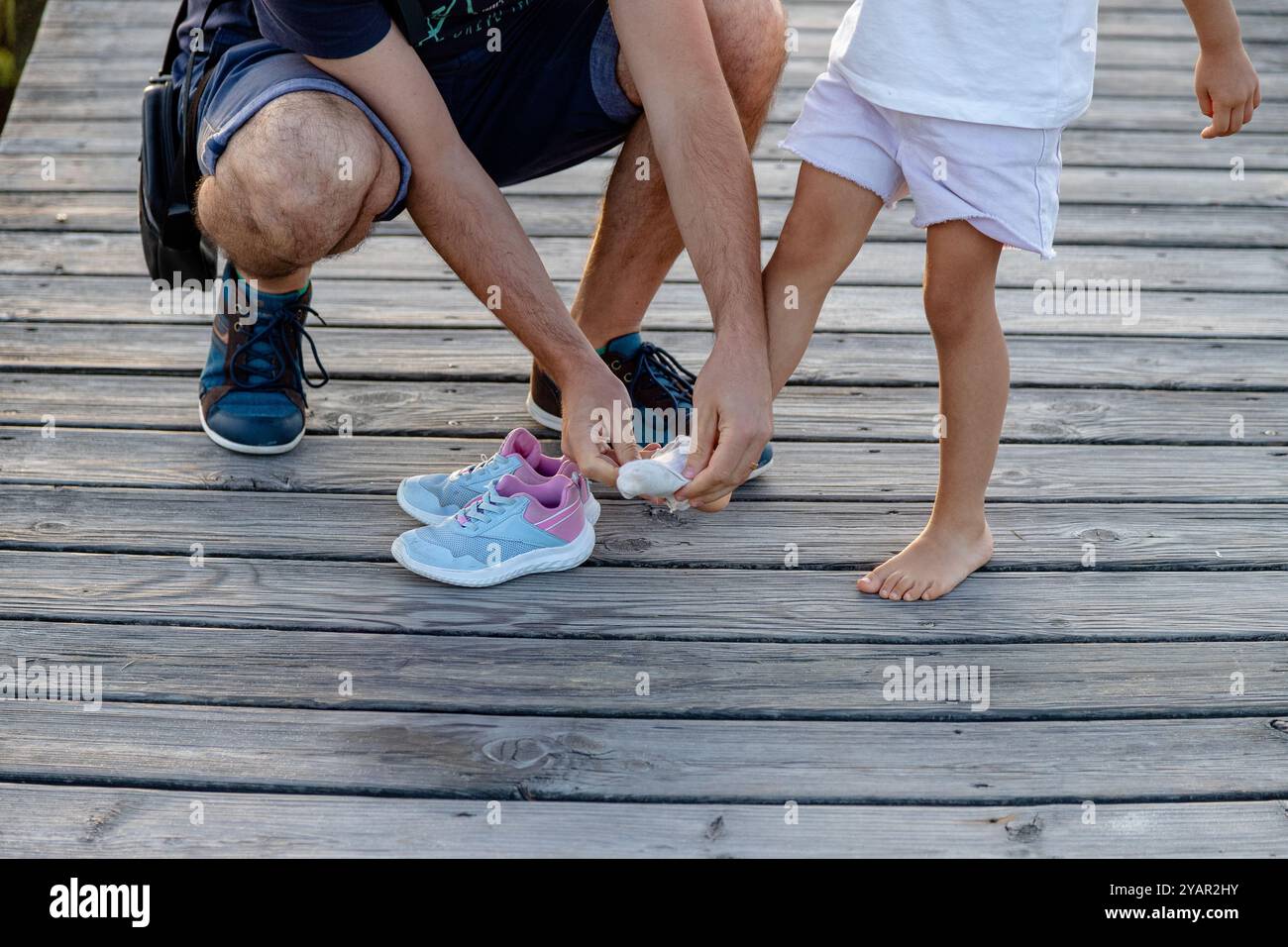 Vater hilft dem Kind, Socken und Schuhe anzuziehen, nachdem er barfuß auf einem Holzsteg gespielt hat. Die Beziehung zwischen Vater und Tochter Stockfoto