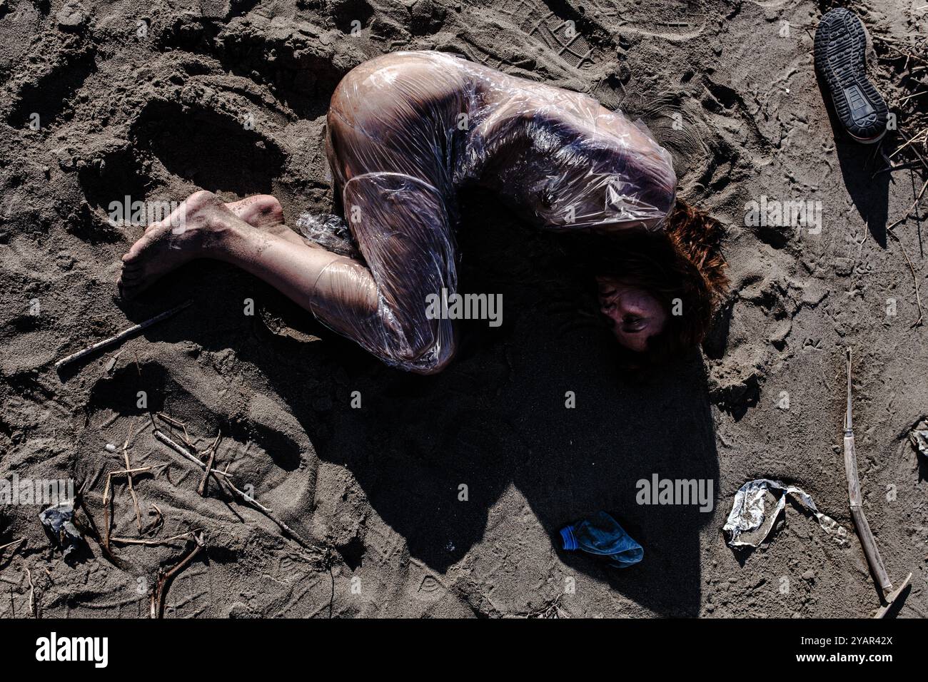 Ein dramatisches und zum Nachdenken anregendes Bild, das eine Person zeigt, die in klares Plastik umhüllt ist und an einem Sandstrand liegt, umgeben von Trümmern. Stockfoto