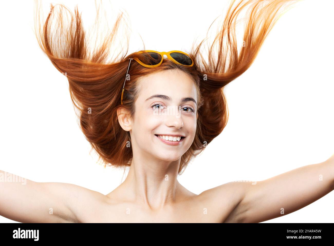 Eine junge Frau lächelt fröhlich mit ihrem leuchtenden, dunkelbraunen Haar, das in der Luft fließt, und trägt eine stilvolle Sonnenbrille auf weißem Hintergrund. Stockfoto