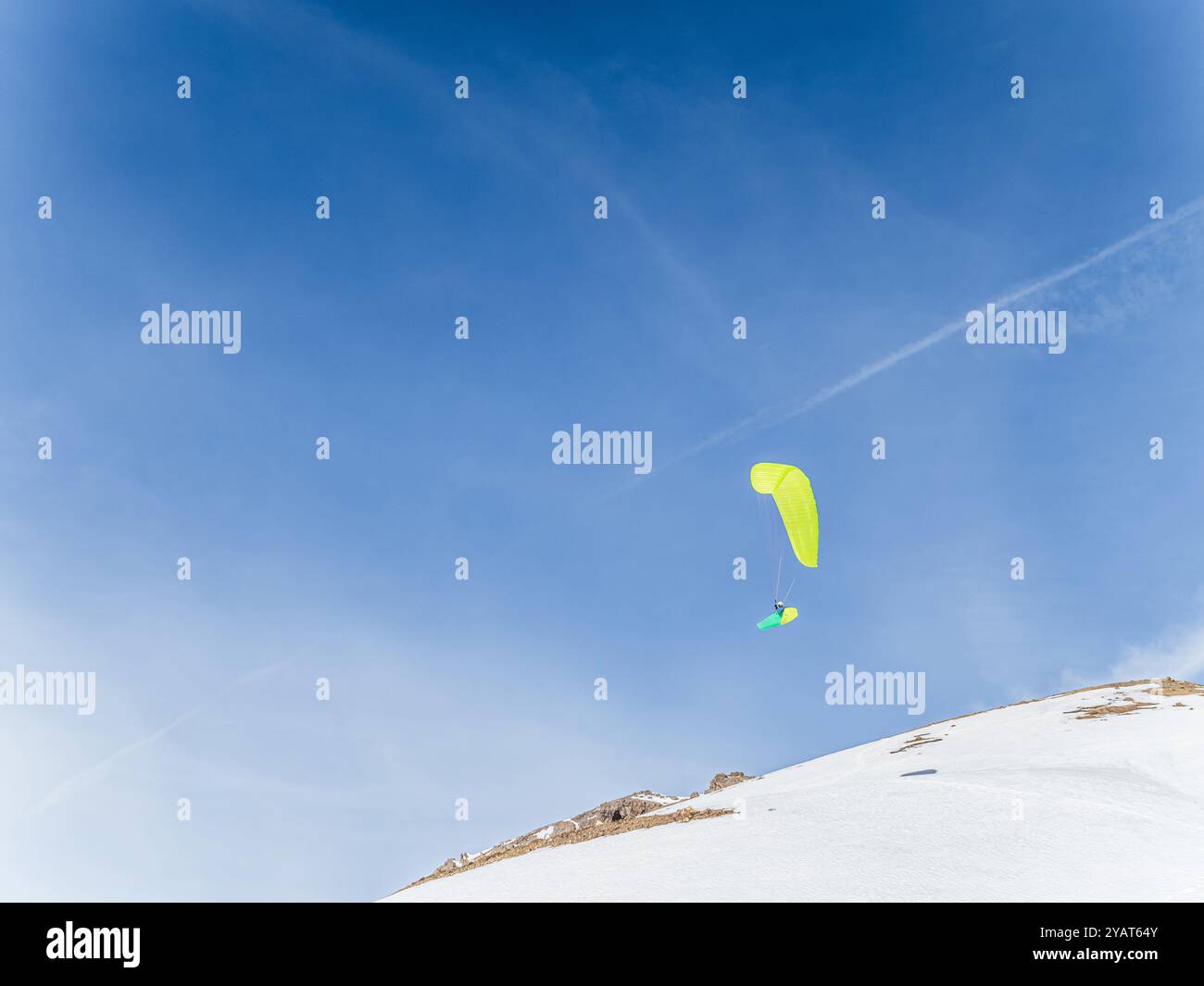 Ein Gleitschirmflieger mit leuchtendem orangefarbenen Flügel schwingt über schneebedeckte Berge in Serre Chevalier, Briancoon, Frankreich. Der helle Himmel und die schroffen Gipfel schaffen das Stockfoto