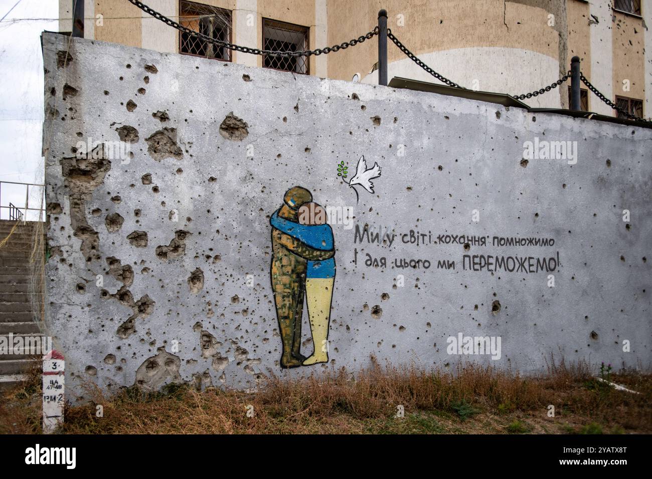 Kupjansk, Ukraine. Oktober 2024. Das Kunstwerk an einer Wand mit Schusslöchern in Kupjansk zeigt eine Person in Militärkleidung, die einen ukrainischen Zivilisten umarmt, mit einem Text, der besagt: „Lasst uns die Welt der Liebe multiplizieren, dafür werden wir GEWINNEN!“. Quelle: SOPA Images Limited/Alamy Live News Stockfoto