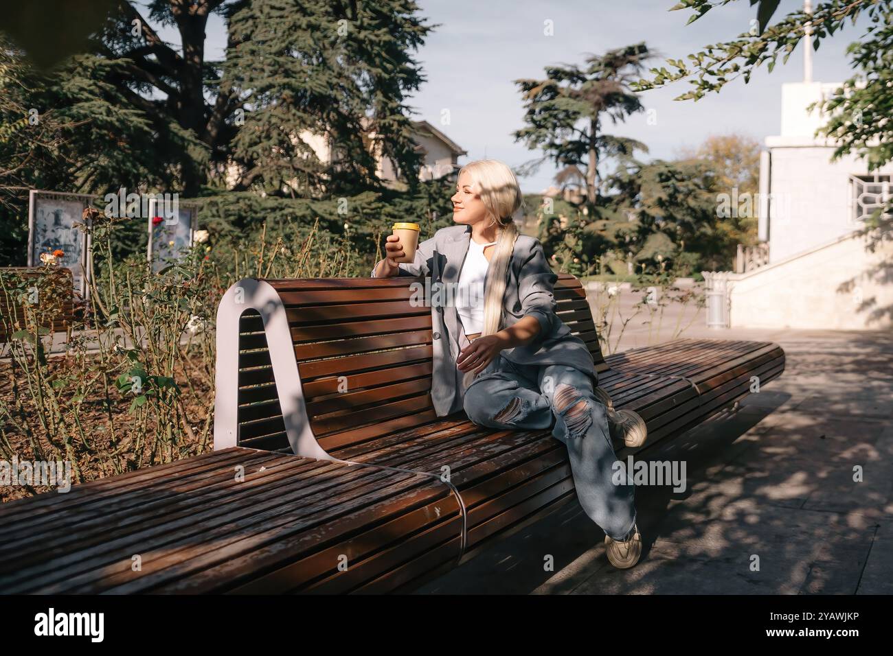 Eine blonde Frau sitzt auf einer Bank mit einer Tasse Kaffee in der Hand. Sie trägt eine graue Jacke und Jeans. Die Bank ist aus Holz und hat einen geschwungenen Rücken Stockfoto