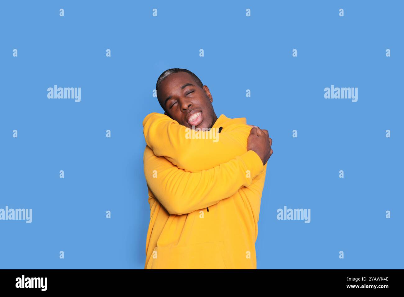 Ein fröhlicher Schwarzer mit einem knallgelben Hoodie umarmt sich mit einem großen Lächeln vor einem leuchtend blauen Hintergrund, der Glück und Kommen ausstrahlt Stockfoto