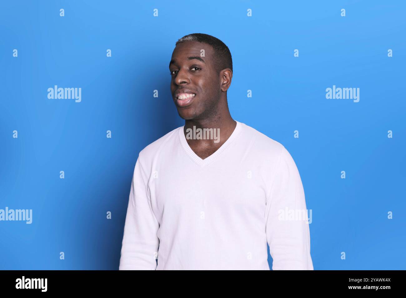 Ein selbstbewusster Schwarzer in einem weißen Pullover steht vor einem schlichten blauen Hintergrund und blickt mit einem freundlichen, offenen Ausdruck direkt in die Kamera. Stockfoto