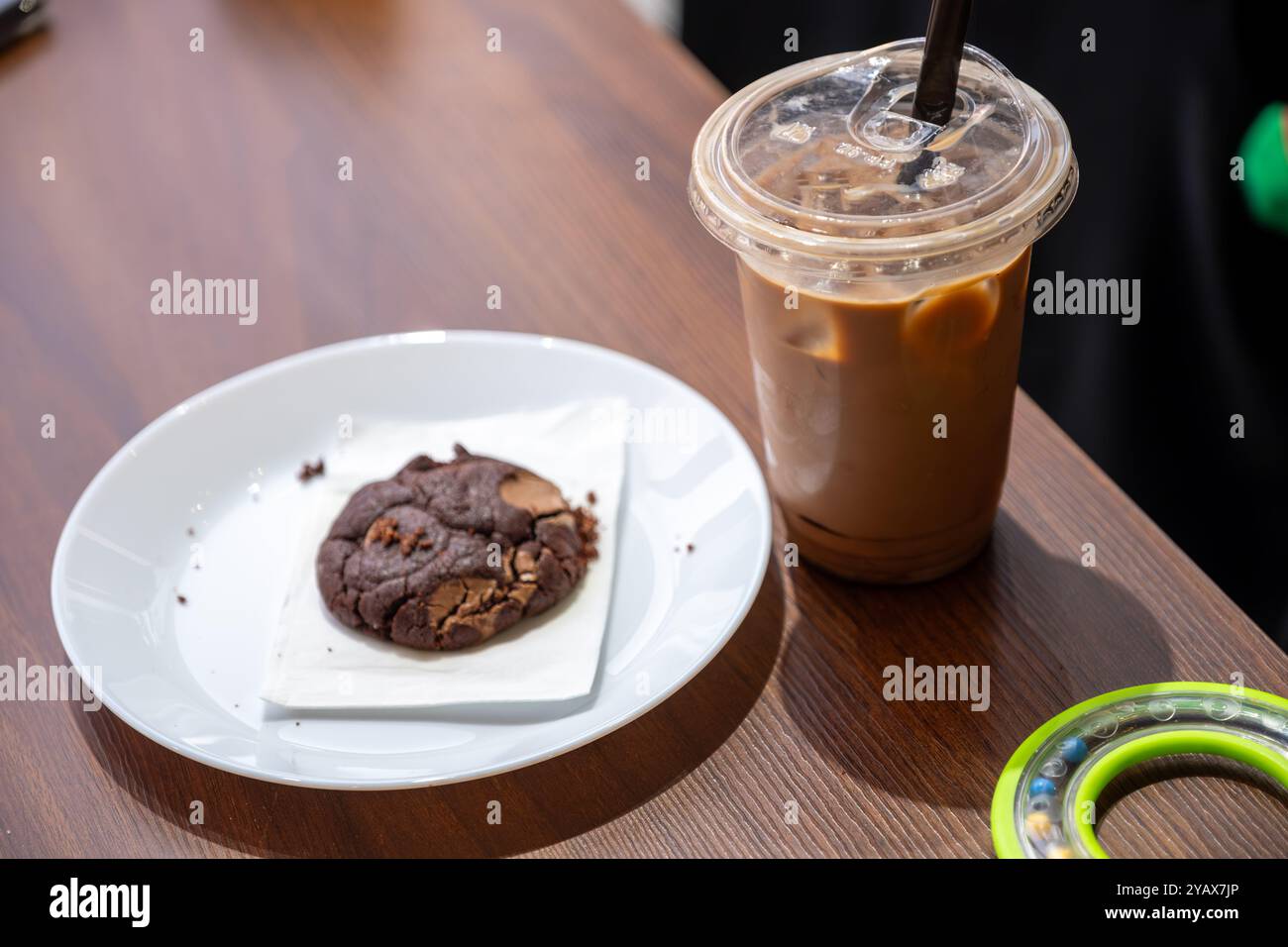 Gekühlter Eiskaffee, serviert in einer Plastiktasse mit schwarzem Strohhalm, begleitet von einem Schokoladenkeks auf einem weißen Teller. Perfekt für eine gemütliche Kaffeepause Stockfoto