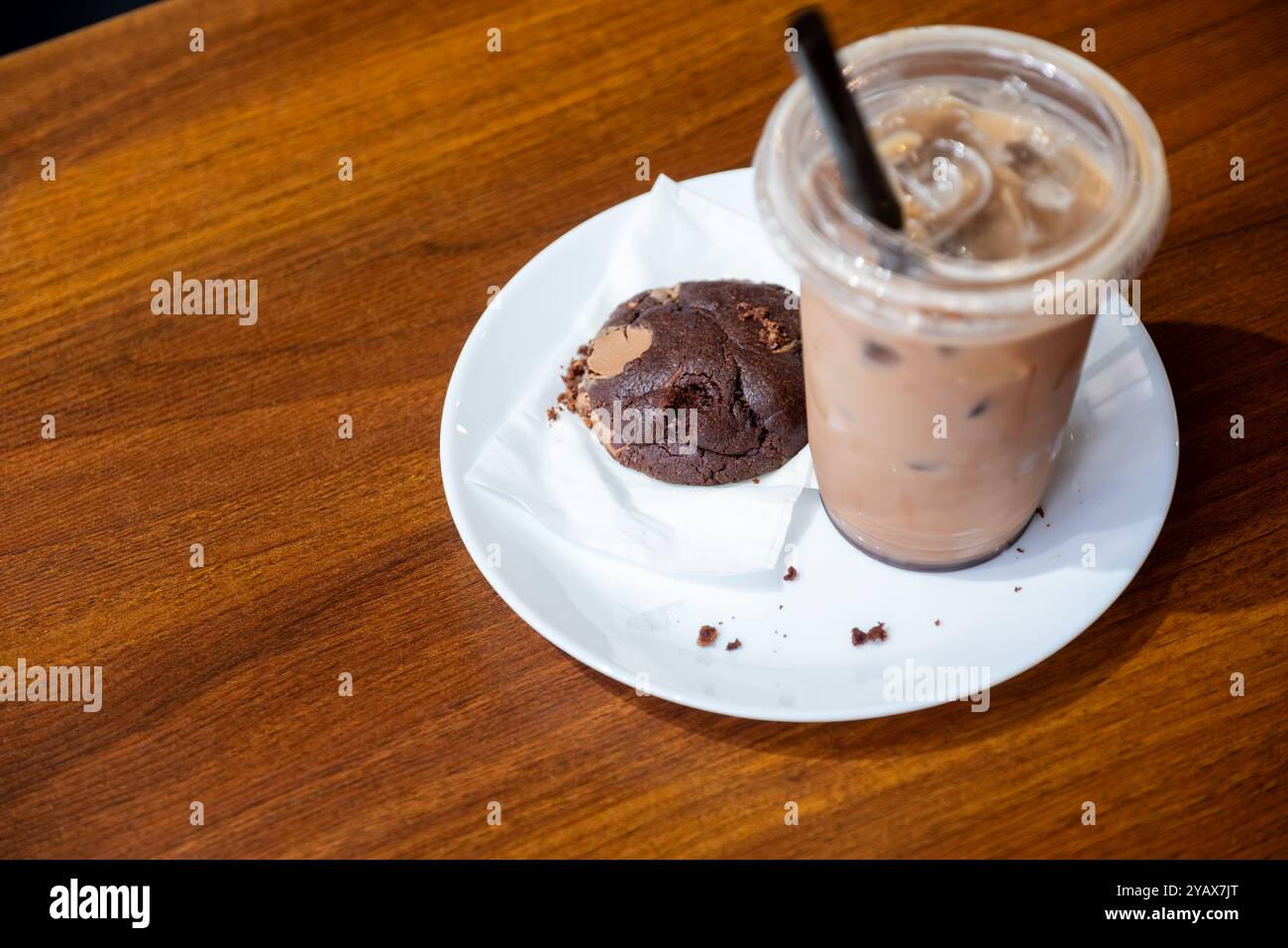 Gekühlter Eiskaffee, serviert in einer Plastiktasse mit schwarzem Strohhalm, begleitet von einem Schokoladenkeks auf einem weißen Teller. Perfekt für eine gemütliche Kaffeepause Stockfoto