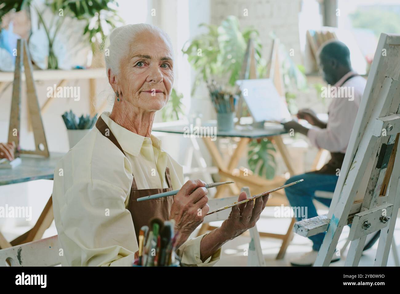 Ältere Frau, die Palette mit Ölfarben und Pinsel hält, während sie im Zeichenstudio in die Kamera schaut Stockfoto