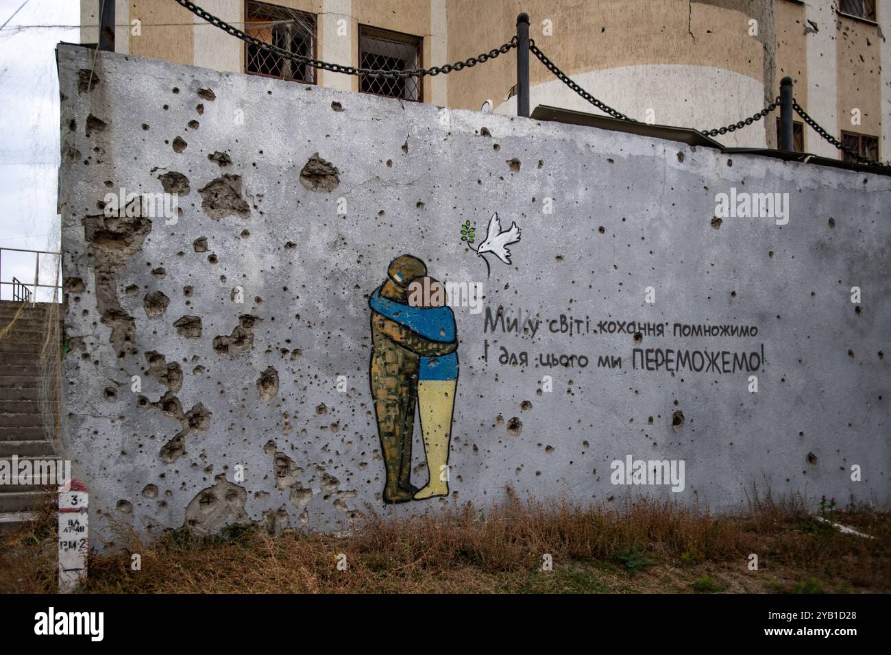 Kupjansk, Ukraine, 13. Oktober 2024. Eine Mauer in Kupjansk, gespickt mit Einschusslöchern, zeigt das Kunstwerk eines Soldaten, der einen ukrainischen Zivilisten umarmt, während eine Friedentaube über ihnen fliegt, mit Text, der locker übersetzt wird: „Lasst uns die Welt der Liebe multiplizieren, dafür werden wir GEWINNEN!“. Quelle: Jay Kogler/Alamy Live News. Stockfoto