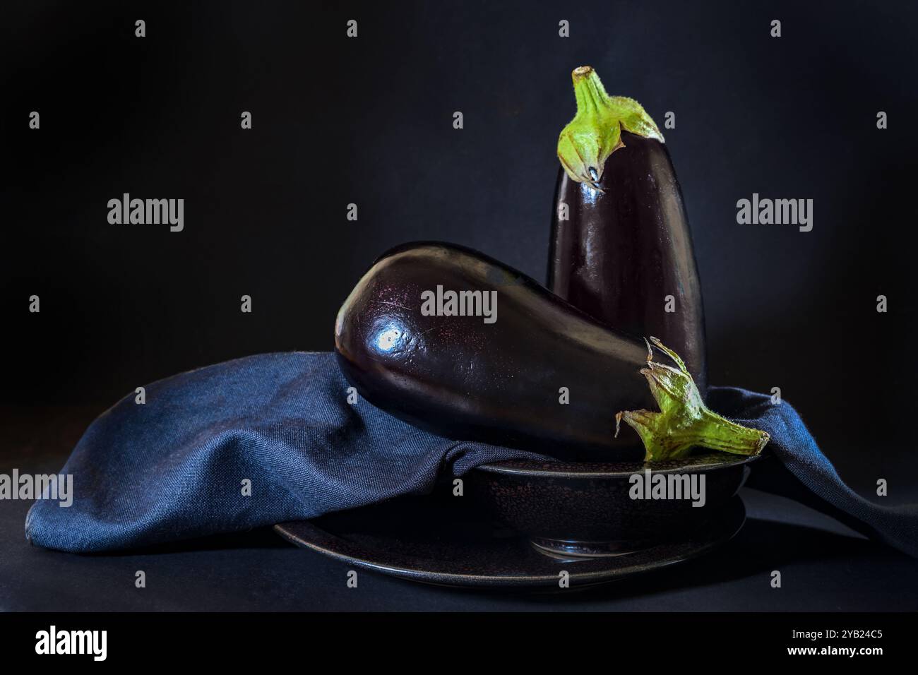 Zwei rohe Auberginen (Solanum melongena) elegant auf einer blauen Serviette in einer Schüssel vor dunklem Hintergrund angeordnet, gesunde Gemüsezutat zum Kochen Stockfoto