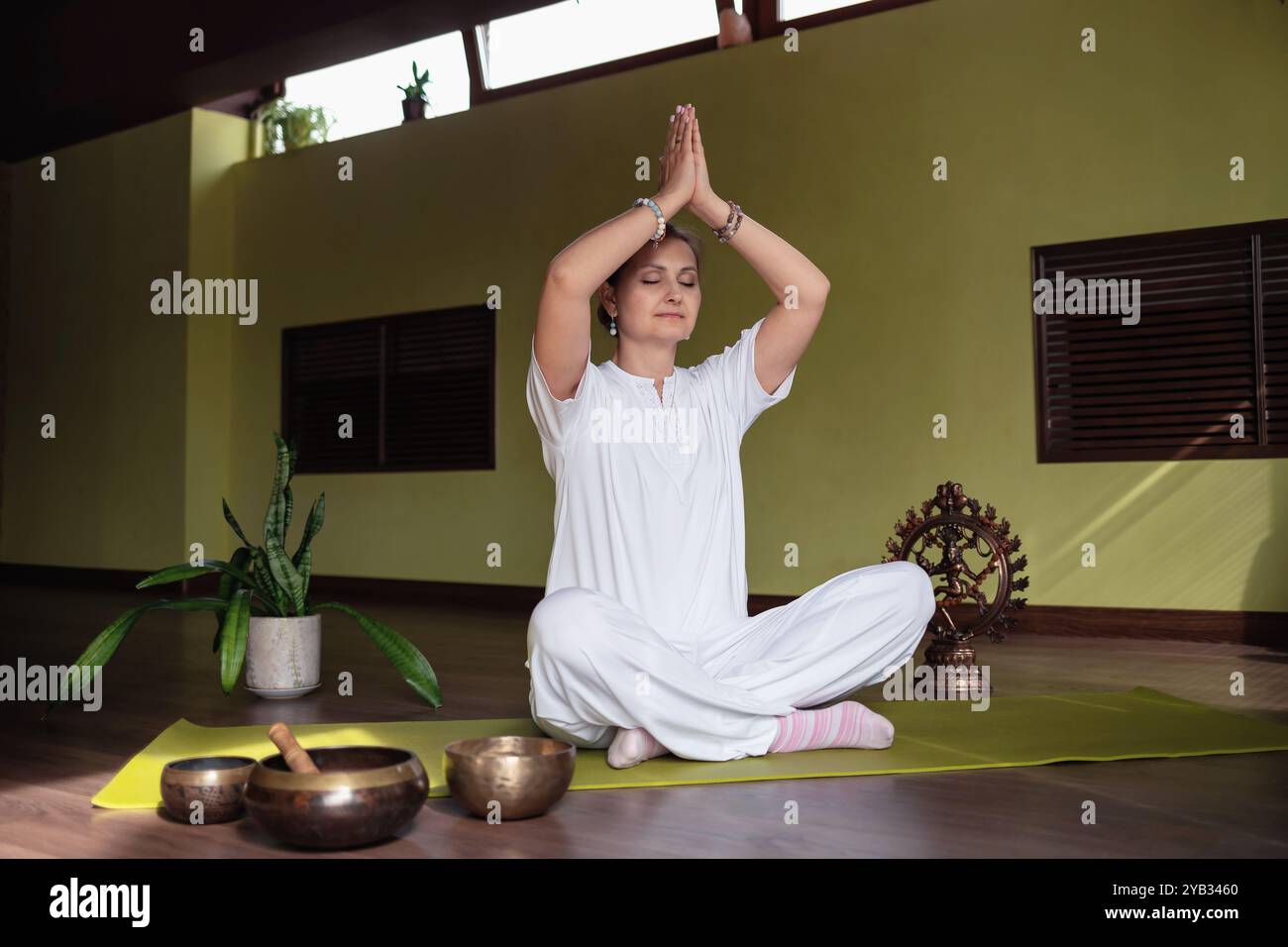 Frau übt Meditation im Kundalini Yoga mit einladender Namaste Geste und erhobenen Händen, sitzt in bequemer Lotusposition auf der Matte im Zimmer Stockfoto