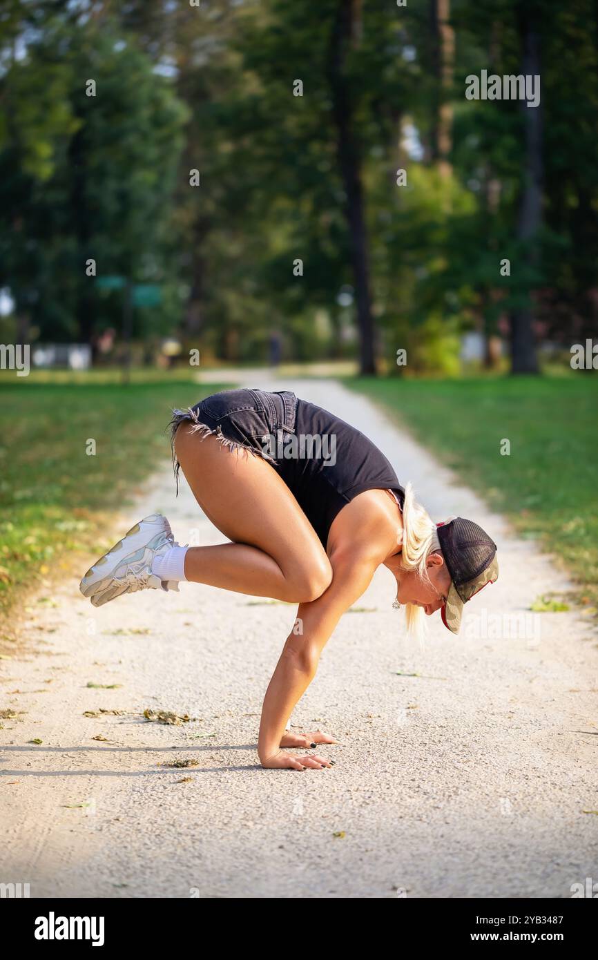 Frau, die Handstand macht, ein Balancierendes Asana namens Bakasana im Yoga, Kranhosen, in lässiger Kleidung auf einem Wanderweg in einem Park üben Stockfoto