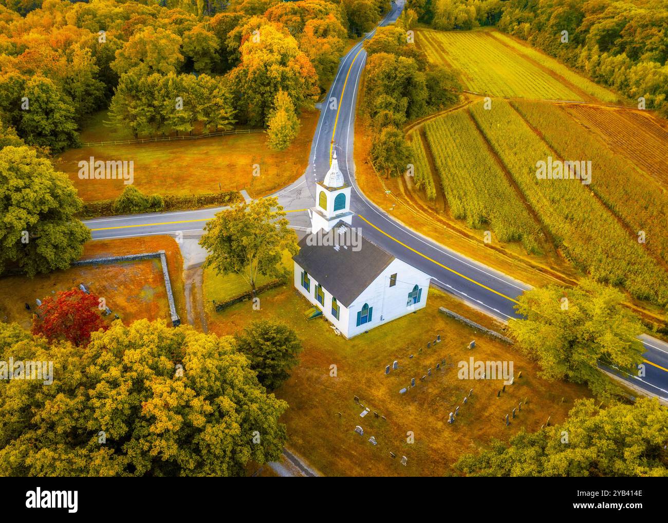 Aus der Vogelperspektive sehen Sie eine malerische Kirche mit einem umliegenden Friedhof, der von einer malerischen, gewundenen Straße durchquert wird. Die Landschaft zeigt einen lebhaften Herbstfol Stockfoto