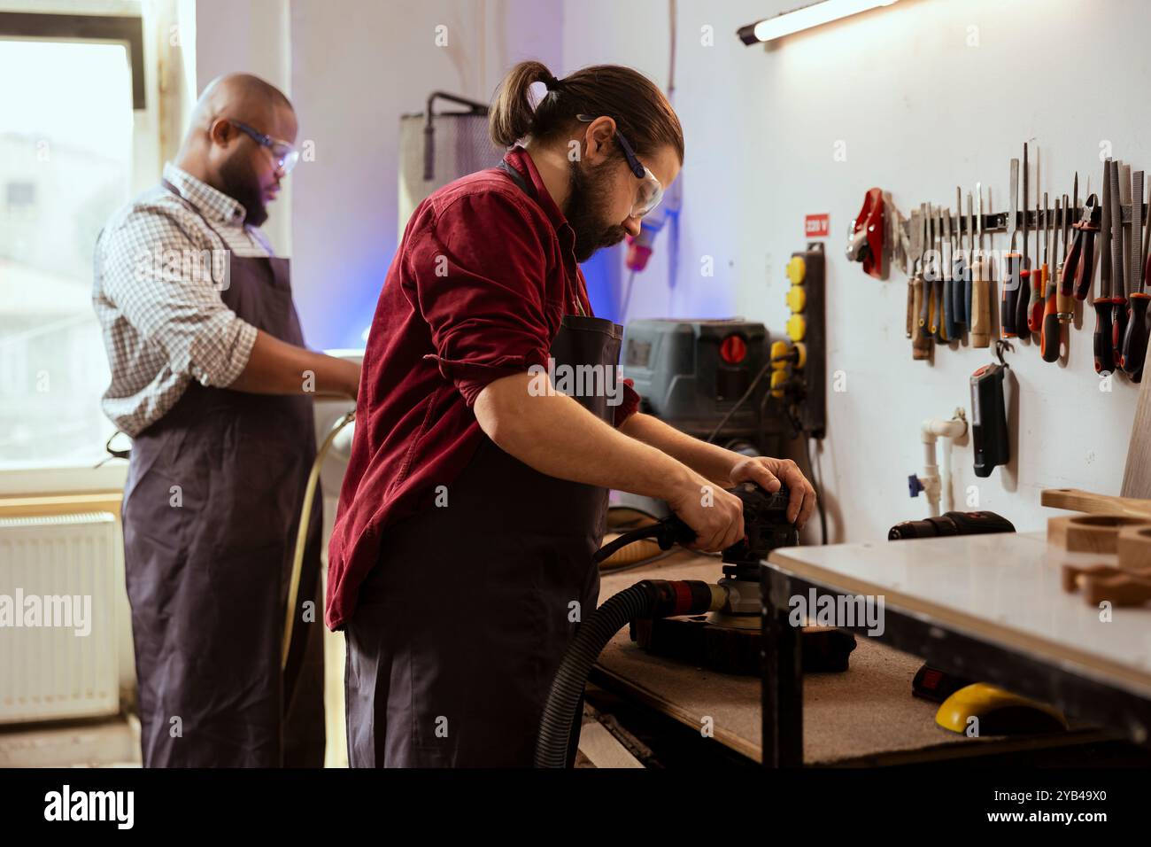 Zimmermann und BIPOC-Mitarbeiter mit Winkelschleifer auf Holzblock, um Oberflächen durch Abrieb mit Schleifpapier zu glätten. Man verwendet Schwingschleifer für die Möbelmontage, unterstützt von einem Kollegen im Hintergrund Stockfoto