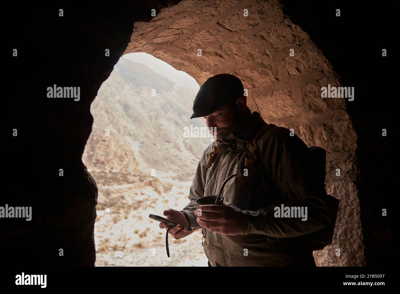 Junge Reisende, die in einem bogenförmigen Fenster eines Zugtunnels stehen, in der einen Hand das Telefon halten, das er benutzt, in der anderen ein Kumpel, ein traditioneller Argentinier Stockfoto