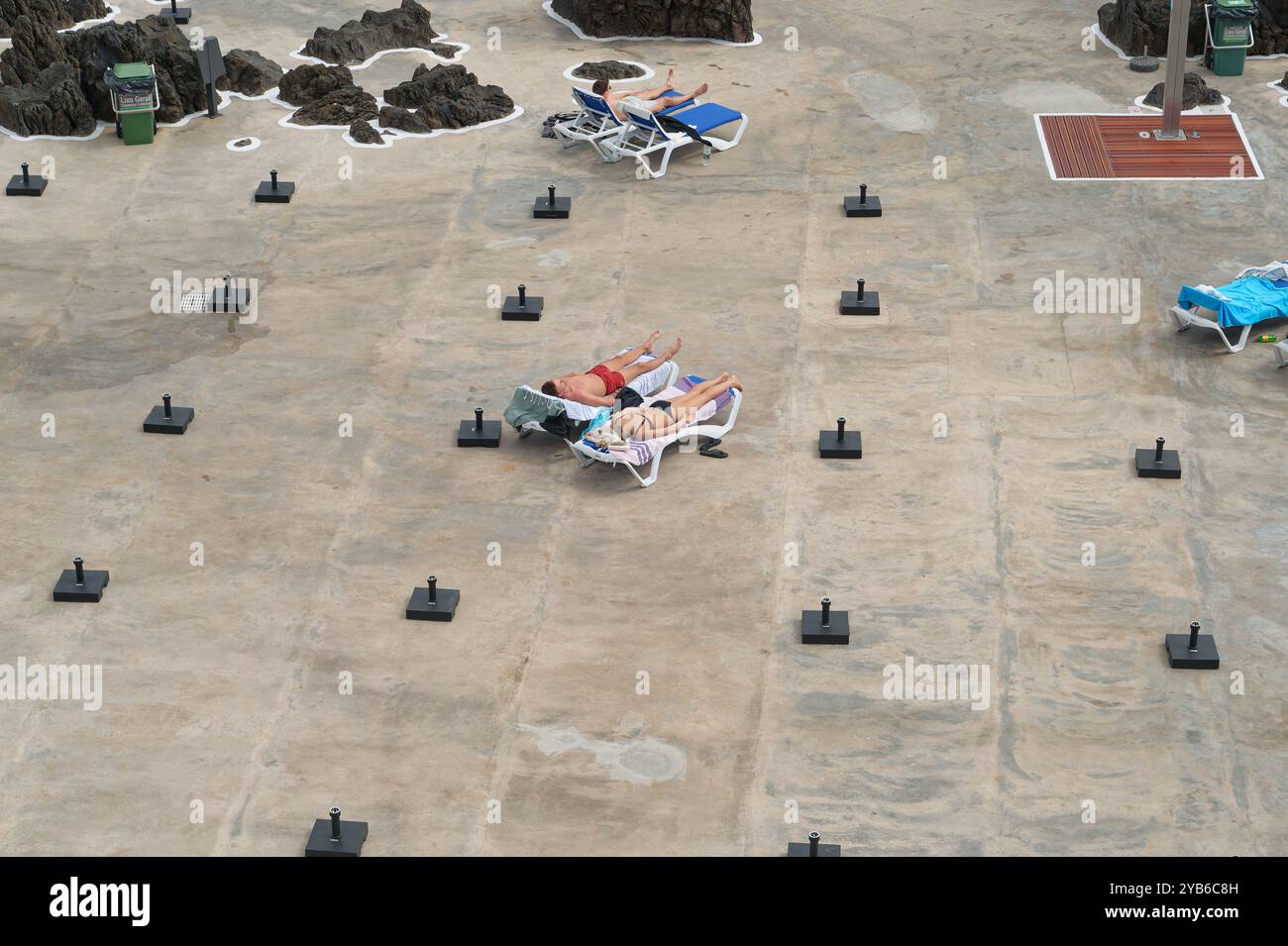 Entspannungsreisende entspannen sich auf Liegestühlen, die von leeren Sonnenschirmen an den natürlichen Pools von Porto Moniz umgeben sind Stockfoto