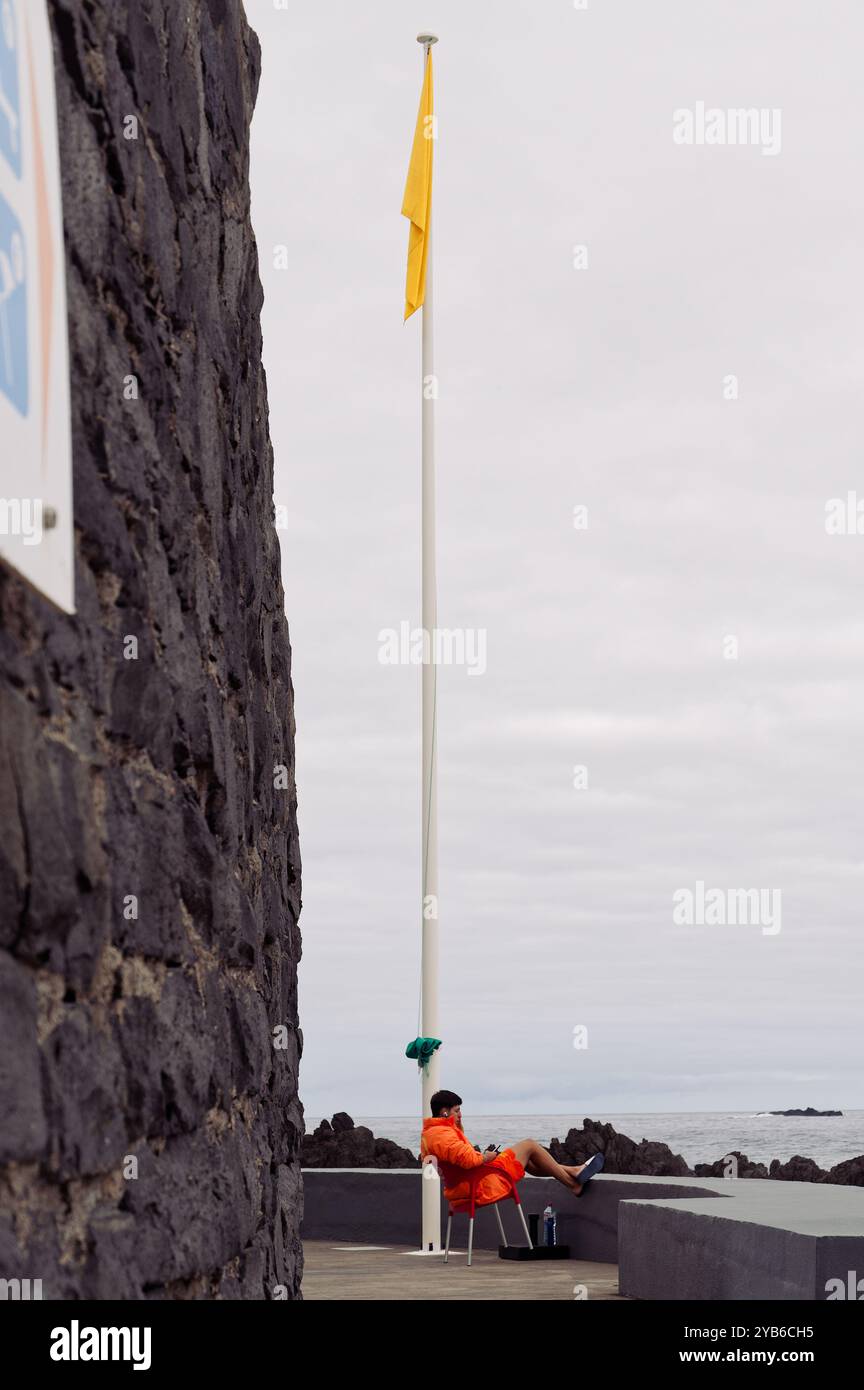 Der Rettungsschwimmer in leuchtendem Orange sitzt unter einer gelben Flagge an den natürlichen Pools von Porto Moniz, neben vulkanischen Steinmauern Stockfoto