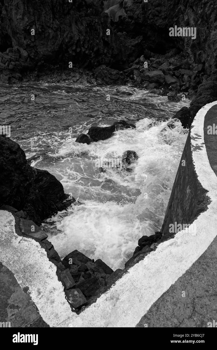 Die Wellen brechen an den natürlichen Pools von Porto Moniz in Schwarz-weiß gegen vulkanische Felsen unter einem scharfen Betonrand Stockfoto