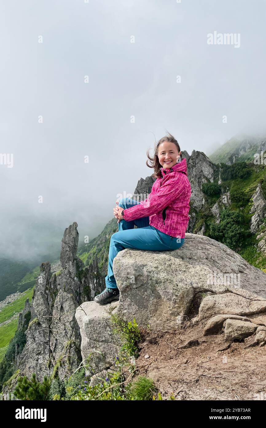 Fröhlich lächelnder Wanderer in rosa Jacke und blauer Hose sitzt auf einem großen Felsen am Rand der Klippe. Hinter ihr sind zerklüftete Felsformationen und grüne Hänge teilweise in Nebel verhüllt. Stockfoto