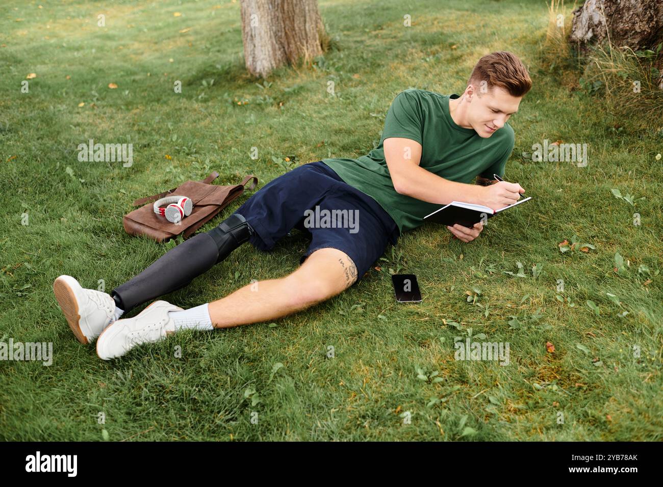 Ein junger Mann mit einer Beinprothese sitzt auf Gras und skizziert in einem Notizbuch, während er entspannt und lächelnd ist. Stockfoto